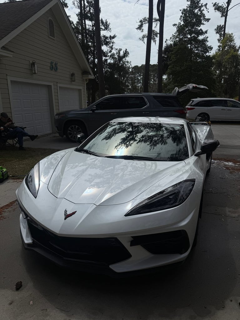 White Chevrolet Corvette parked in a driveway with a garage and other vehicles visible in the background