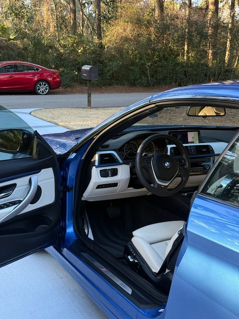 Blue BMW coupe with open driver door showing white and black interior, parked in driveway with red car and forest background