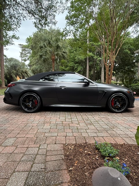 Black convertible sports car parked on brick driveway with trees and landscaping in background