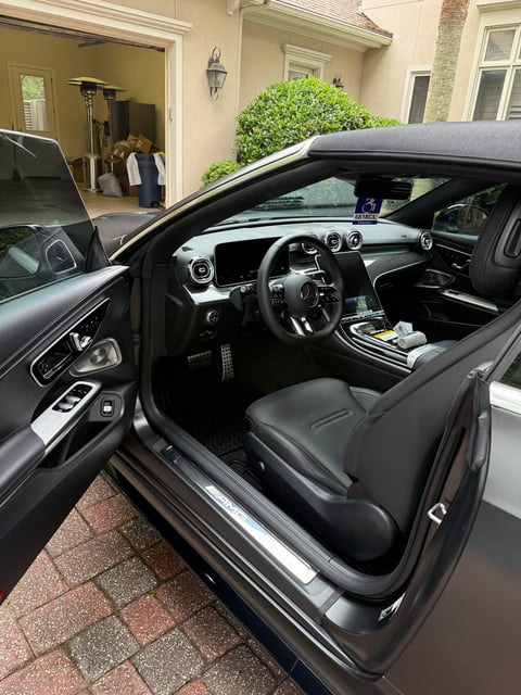 Interior of a black sports car with open driver door, showing dark leather seats and steering wheel, parked in a driveway with house visible