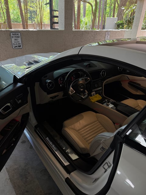 Interior view of a white convertible sports car with tan leather seats and dark steering wheel in a showroom with glass windows overlooking trees