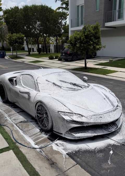 White sports car covered in white foam during washing on suburban street with trees and houses