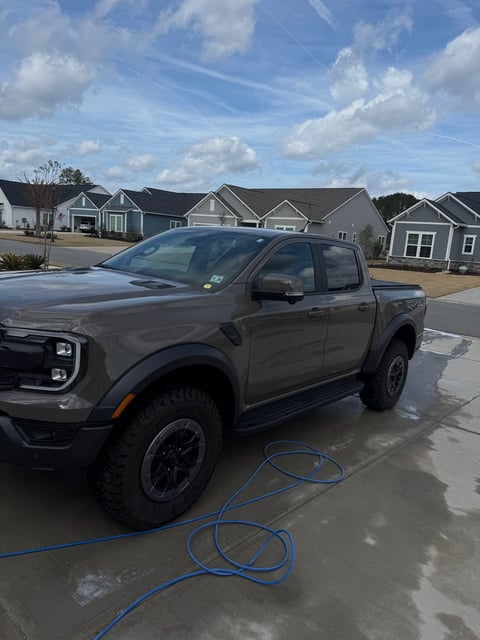 Gray pickup truck parked in driveway with blue hose, suburban residential homes in background under blue sky