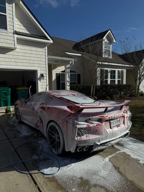 Red sports car covered in soap suds during outdoor washing in residential driveway