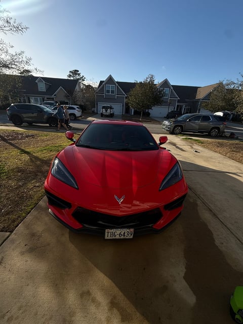 Red Corvette sports car parked on a residential driveway with suburban houses and other vehicles in the background