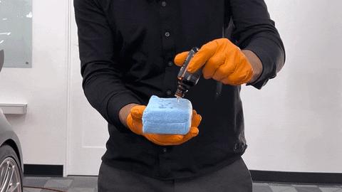 Person in black clothing and orange gloves holding a blue soapy sponge and spray bottle for car washing